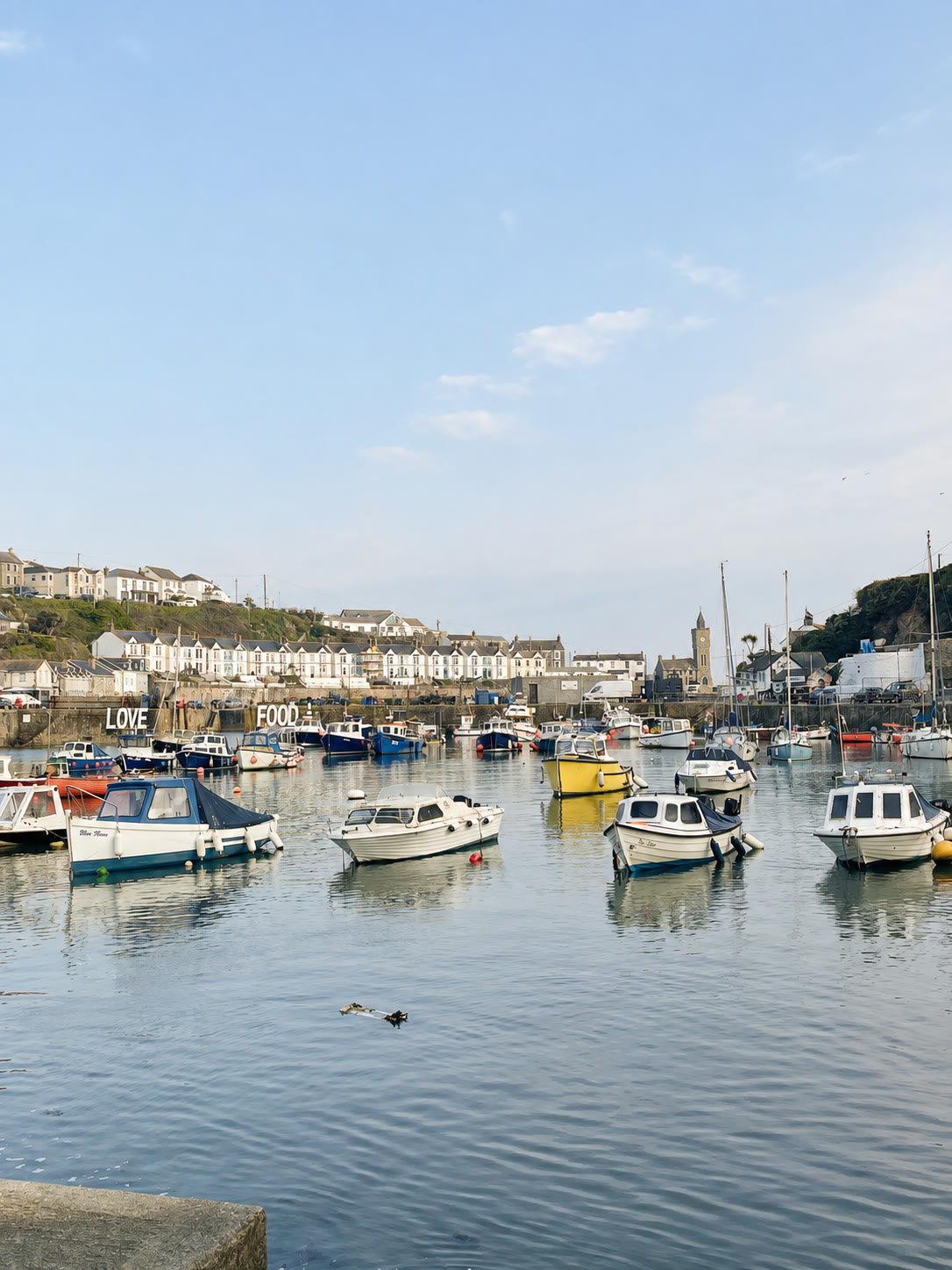 Boats on the blue water of Porthleven harbour, with the clock tower and Cornish houses beyond.